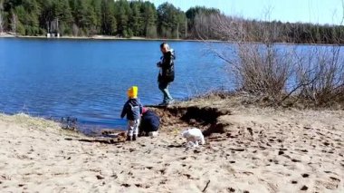 Küçük bir köpek sakin bir göl kenarında kıyı şeridini keşfederken iki çocuk kumdan kaleler inşa ediyor. Bir kadın onları uzaktan izliyor..