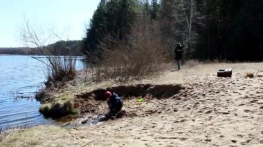 İki çocuk sakin bir gölün yanındaki kumlu bir plajda oyuncu aktivitelere girişiyor. Bir çocuk suları incelerken diğeri açık mavi gökyüzünün altında doğal ortamın tadını çıkarıyor..