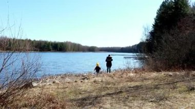 Bir çocuk ve bir yetişkin sakin bir gölün kenarında yürüyor, güneşli havanın ve doğanın huzurunun tadını çıkarıyorlar. Arka planda yemyeşil ağaçlar var. Huzurlu atmosfere katkı sağlıyor..