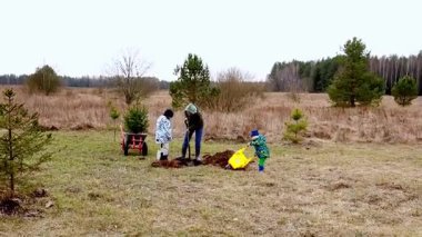 Yeşilliklerle dolu bir düzlükte bir aile ağaç dikiyor. İki çocuk, birlikte kaliteli zaman geçirirken toprağı kazmak ve hazırlamak için aletler kullanarak ebeveynlerine yardım ediyor..