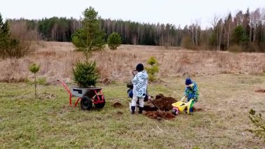 İki çocuk açık bir alanda kürek ve el arabası kullanarak küçük ağaçlar dikmekle meşgul. Sahne huzurlu, ağaçlar uzakta ve bulutlu bir gökyüzü tepemizde..