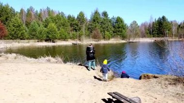 Bir aile sakin bir gölün kenarında güneşli bir bahar gününün tadını çıkarır. Çocuklar suyun kenarında oynarken, yetişkinler seyrediyor. Bereketli yeşillik huzurlu ortamı sarar, neşeli bir atmosfer yaratır..