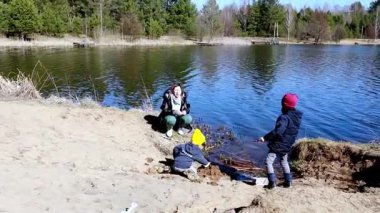 İki çocuk suyun yakınındaki kumu kazmaktan zevk alırken bir kadın onları yakından izliyor. Sahne güneşli bir günde ağaçlarla çevrili pitoresk bir göl, açık hava eğlencesi için mükemmel..