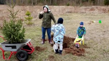Bir aile bir tarlaya genç ağaçlar dikmekle meşgul. İki çocuk kürek ve el arabasında yardımcı oluyor, serin bir günde açık havada eğleniyorlar..