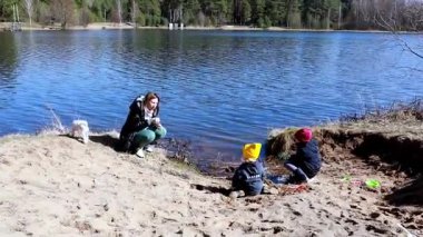 İki çocuk sakin bir gölün yakınındaki kumda oynarken anneleri onları denetliyor. Yakınlardaki bir köpek, ılık bir bahar gününde neşeli bir atmosfere katılır..