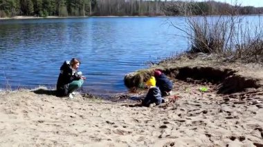 İki çocuk göl kenarında kumu kazarken bir yetişkin onları izliyor ve onlarla etkileşime geçiyor. Aydınlık bir gün ve çevre sakin ve doğal..