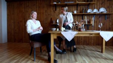 İki kadın ahşap bir masada oturmuş, sıcacık, kırsal bir yemek alanında sohbet ediyorlar. Ortam sıcak, masa ayarları ve hafif ışıklandırma deneyimi güçlendiriyor..
