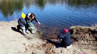 İki çocuk, yanında bir yetişkin otururken sakin bir su kütlesinin yanında kumda oynuyor. Güneşli bir gün, açık hava maceraları için mükemmel bir zemin sağlar..