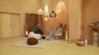 İki kadın, eklektik dekorlarla dolu sıcak, davetkar bir odada rahatlar. Yumuşak ışıklandırma ve rahat döşemelerle çevrili iken canlı bir sohbete dalarlar ve sakin bir atmosfer yaratırlar..