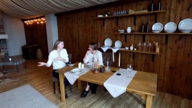 İki kadın, sıcak ve davetkar bir atmosferle çevrili ahşap bir masada otururken sohbet ediyorlar. Ayar, mumlar ve sofra takımlarıyla güzel düzenlenmiş bir masa içeriyor..