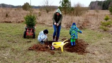 Aileler kırsal bir alanda toplanıp ağaç dikiyorlar. İki çocuk çukur kazarken bir yetişkin de genç fidanları dikmek için hazırlıyor..