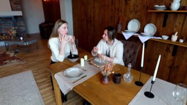 İki kadın, nefis sofra takımları ve ortam aydınlatmalarıyla dolu, güzel, ahşap bir ortamda, yemek masasında canlı bir tartışmayı paylaşıyor, davetkar bir atmosfer yaratıyor..