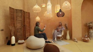İki kadın sıcak renklerle, dokunmuş dekorlarla ve yumuşak ışıklandırmayla süslenmiş rahat bir odada düşünceli bir tartışmaya girişirler. Bağlanmak için mükemmel bir ortam yaratırlar..