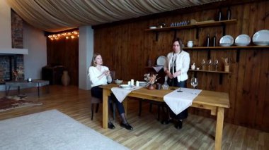 İki birey güzel tasarlanmış kırsal bir iç mekanda zevkli bir sofra dekoruyla davetkar bir yemek atmosferi yaratmak için işbirliği yapıyor. Ayarda ahşap elementler ve sıcak ışıklandırma yer alıyor.