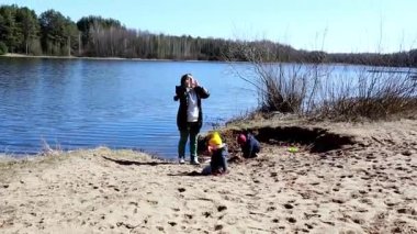 Bir anne, sakin bir gölün kenarında neşeyle durup güneşi emerken, iki küçük çocuğu da yakınlardaki kumlu bölgede oyun oynuyor. Arkaplanda ağaçlar ve açık mavi gökyüzü yer alıyor..