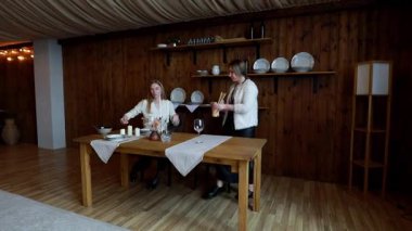 Sıcak, ahşap bir alanda, iki kadın basit ama zarif ayarları olan bir yemek masası hazırlarlar. Çiçekler, cam eşyalar ve masa örtüleri rahat atmosfere cazibe katar..