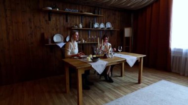 İki kişi, sıcak ve kırsal bir odada yemek yemek için hazırlanan ahşap bir masada oturuyor. Yumuşak ışık pencereden süzülerek yemek için rahat bir atmosfer yaratır..