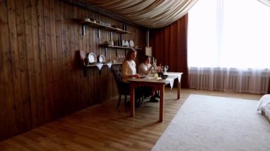 İki kişi, gündüz saatlerinde sıcak ahşap bir iç mekanda bulaşıkları olan düzenli bir masada sohbet ediyorlar. Sıcaklık davetkar atmosferden yayılır..