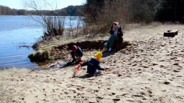 Çocuklar deniz kenarındaki kumda oynarken yetişkinler güneşli bir günde dinleniyorlar. Huzurlu göl manzarası ağaçlarla çevrili. Aileler için huzurlu bir ortam yaratıyor..