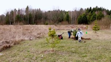 Ebeveynler ve çocuklar birlikte çalışır, yeşilliklerle çevrili açık bir alana yeni fidanlar ekerler. Doğayı beslerken atmosfer neşe dolu..