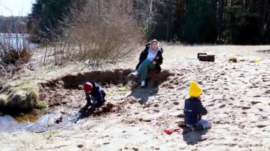 Çocuklar göl kenarındaki kumlu sahili kazıp keşfederken ebeveynleri yakınlarda dinleniyor. Sahne, bahar güneşinde açık havada oynamanın neşesini ve yaratıcılığını yakalıyor..