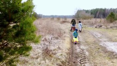 Bir aile huzurlu bir kırsalda geziniyor, çocuklar mutlu bir şekilde toprak bir patika boyunca yük arabası çekiyor. Manzara, çıplak ağaçlar ve açık arazilerle huzur dolu..