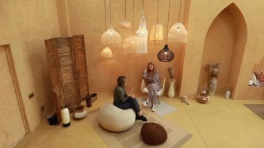 Sıcak ve davetkar bir mekanda, iki kadın rahat bir şekilde yumuşak koltukta oturur, güzel bir şekilde hazırlanmış ışıklandırma ve dekorasyonla çevrili sohbetin derinliklerinde. Atmosfer yaratıcılık ve sıcaklık yayıyor..