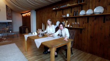 İki kadın, sıcak, kırsal bir mekanda, mum ve yemek takımları yerleştirerek zarif bir yemek masası hazırlıyor. Atmosfer davetkar, samimi ve misafirperver..