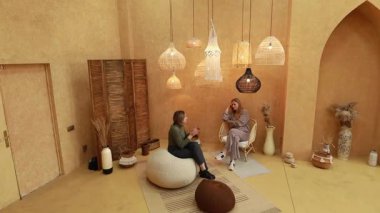 İki kadın, çeşitli lamba armatürlerinin yer aldığı sıcak, davetkar bir odada oturuyorlar. Dekoratif elementlerle çevrili konuşmalarla meşgul olduklarında atmosfer rahatlıyor..