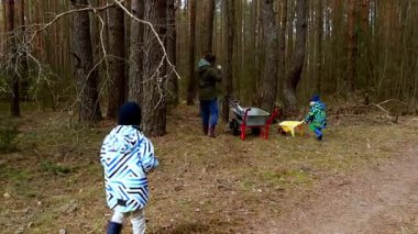 İki genç çocuk, orman ortamında bir yetişkine neşeyle yardım ediyor, küçük bir arabayı çekiyor ve ağaçlık bir yolda el arabasını itiyor. Uzun ağaçlarla çevrili açık hava aktiviteleriyle meşguller..