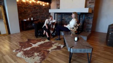 İki kadın kırsal bir salda oturmuş keyifli bir sohbete dalıyorlar. Dekorasyonda bir şömine ve ahşap bir dekor var. Rahatlamak için mükemmel bir ortam yaratıyor..