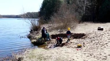 Bir aile göl kenarında güneşli bir gün geçirir. Kumda oynayan bir çocuk ve ağaçlarla çevrili bir kumdan kale yapımına yardım eden bir yetişkin..