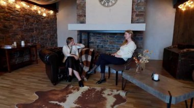 İki arkadaş, şık bir salonda rahat bir sohbete girişiyor. İçinde taş bir duvar, yumuşak mobilyalar ve sıcak bir aydınlatma var. Hoş bir atmosfer yaratıyor..