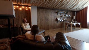 Ahşap duvarlarla süslenmiş rahat bir oturma odasında, iki kadın rahatça oturur. Yumuşak ışıklandırma ve zevkli dekorlarla çevrili bir ortamda sıcak bir atmosfer yaratarak sohbet ediyorlar..