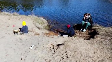 İki çocuk nehir kenarındaki kumda oynuyor, ebeveynleri yakınlarda dinlenirken oyuncaklarla inşa ediyorlar. Sahne güneşli güzel bir gün yakalıyor, açık hava aktiviteleri için mükemmel..