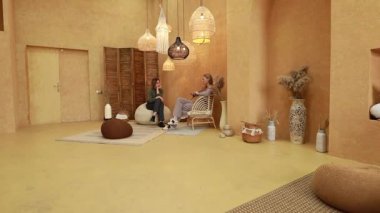 İki birey, benzersiz ışıklandırma ve doğal dekorasyonlar içeren ılık tonlu bir iç mekanda sakin bir sohbete girişiyor. Atmosfer davetkar ve huzur dolu..