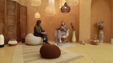 İki kadın, sıcak tonlarla ve doğal malzemelerle dekore edilmiş şık bir odada oturmuş, rahat ve davetkar bir atmosferde çeşitli konuları tartışıyorlar..