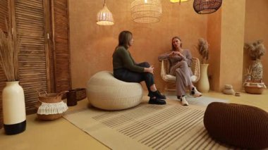 İki kadın, modern tasarım elementleri ve dokularının yer aldığı şık, sıcak dekore edilmiş bir alanda rahat bir şekilde otururken derin bir sohbete girişirler..