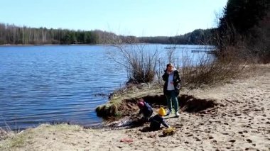 Güneşli bir günde, kumlu bir sahilde oynayan iki çocuk, ağaçlar ve suyla çevrili, kazma ve inşa etme gibi aktivitelere girişiyor..