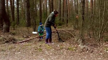 Palto ve bot giymiş bir kadın, bir orman yolundan düşen dalları ve enkazı temizliyor. Çevredeki bölge yüksek ağaçlar ve sonbahar yapraklarıyla dolu. Bu da sakin bir atmosfer yaratıyor..