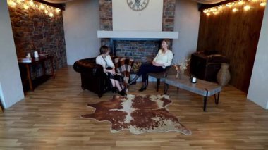 İki kadın rahat bir koltukta otururken derin bir sohbete dalarlar. Etrafı zevkli dekorlar ve sakin bir ortamda hafif aydınlatmalarla çevrilidir..