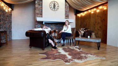 İki arkadaş şık bir oturma odasında rahatça otururlar. Biri koltuğa yaslanırken diğeri rahat bir sohbete daldı. Etrafı kırsal dekor ve hafif ışıklandırmayla çevrili..