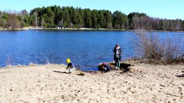 Bir yetişkin fotoğraf çekerken iki çocuk sakin bir gölün yakınındaki kumu kazıyor. Doğayla ve ağaçlarla çevrili güneşli bir bahar gününün tadını çıkarırlar..