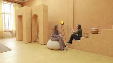 İki arkadaş sıcak, minimalist bir odada oturmuş sohbet ediyorlar. Mekanda topraksı tonlar, basit mobilyalar ve dekoratif elementler var. Rahat bir atmosfer yaratıyorlar..