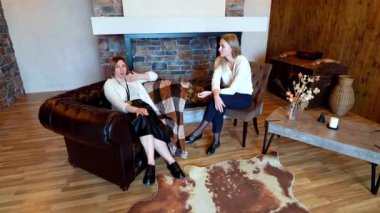 İki kadın, sıcak tonlu mobilyalar ve taş bir şömineyle çevrelenmiş bir şekilde özel hayat hikayelerini ve sezgilerini tartışırken büyüleyici bir yaşam alanında rahat bir şekilde oturuyorlar..