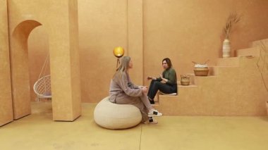 İki kadın minimalist dekorlarla dolu sakin, toprak renkli bir odada oturuyor. Doğal elementler ve yumuşak ışıklandırmayla çevrili anlamlı bir konuşmayı paylaşıyorlar, sakinleştirici bir ortam yaratıyorlar..