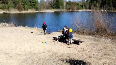 Çocuklar sakin bir göl kenarı ortamında aktif olarak kumdan kaleler inşa ederken, yetişkinler yakınlarda dinleniyor, sıcak güneş ve huzurlu bir çevrede sırılsıklam oluyorlar..