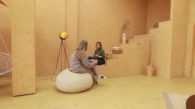 Huzurlu, minimalist bir ortamda, iki arkadaş, yuvarlak bir yastıkta rahatça otururken canlı bir sohbete dalarlar. Sıcak renkler davetkar bir atmosfer yaratır..