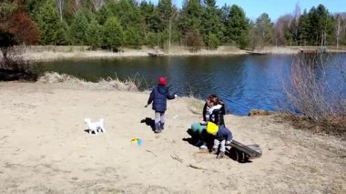 Çocuklar ağaçlarla çevrili sakin bir gölün yanındaki kumlu kıyılarda eğlenceli aktivitelere girişiyor. Birbirleriyle gülüyor ve etkileşime geçiyorlar, dışarıda güzel bir günün tadını çıkarıyorlar..