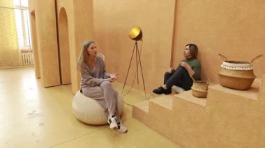 İki kadın, sıcak renkli bir iç mekanda ev dekorları hakkında sohbet ediyorlar. Yumuşak dokular ve rahat mobilyalar fikirleri paylaştıkça davetkar bir atmosfer yaratıyor..